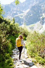 Naklejka na ściany i meble Young woman with hiking equipment walks along a hiking trail against the backdrop of mountain scenery. Summer vacation trip on weekend. Active lifestyle
