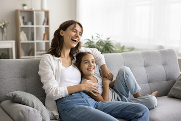 Obraz premium Happy excited young mom and cheerful daughter kid having fun on soft couch at home, relaxing on sofa, looking away, laughing, enjoying family leisure, comfort, relaxation, close relationships