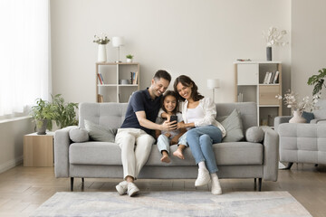 Positive young mom, dad and sweet girl kid taking family selfie on cozy comfortable couch, having...