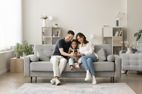 Positive Young Mom, Dad And Sweet Girl Kid Taking Family Selfie On Cozy Comfortable Couch, Having Fun With Digital Gadget In Cozy Stylish Home Interior, Using Smartphone For Selfie Together