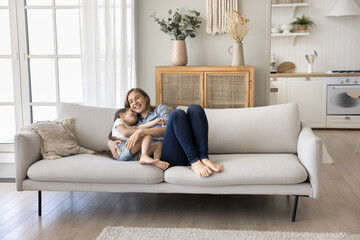 Obraz premium Sweet moments of loving mother and little daughter. Cute preschooler kid girl relaxing on sofa cuddling with loving mom, laughing, play, sit on couch, enjoy carefree rest in cozy living room at home