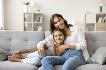 Happy young mom cuddling adorable girl kid, resting on comfortable couch, looking at camera, smiling, laughing. Cheerful little daughter child and mother enjoying family leisure, childhood - obrazy, fototapety, plakaty