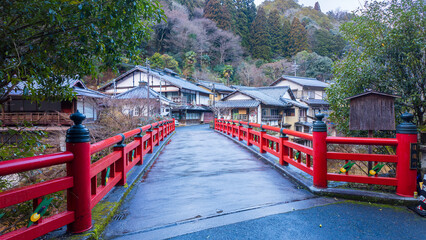 Red traditional Japanese bridge to small village in forested mountains - 712770471
