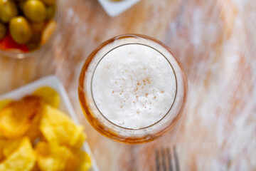Glass of chilled foam frothy beer with snacks - portion of yellow crispy potato chips on small...