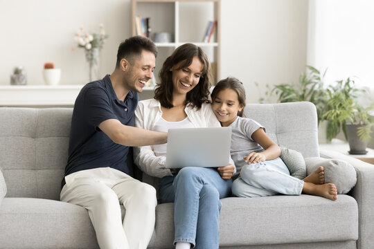 Happy young parents and cute kid relaxing at laptop on sofa, holding computer on lap, using online media service for watching movie, entertainment, enjoying Internet technology, connection at home