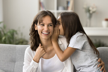 Cheerful positive little girl whispering secret at laughing mothers ear. Happy mom and daughter kid having fun on home sofa, talking, laughing, feeling closeness, trust, enjoying family leisure