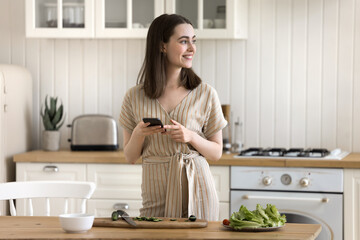 Young pretty woman standing in kitchen prepare vegetarian dish, distracted for cookery, using...