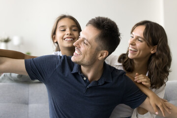 Obraz premium Happy parents piggybacking playful little daughter kid for playing airplane flight, sitting on comfortable home sofa, laughing, smiling, having fun, enjoying parenthood, family time