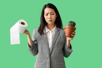 Young Asian businesswoman with cactus and toilet paper on green background. Hemorrhoids concept
