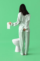 Young woman in pajamas with paper roll and toilet bowl on green background, back view. Hemorrhoids...