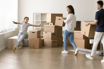 Cheerful young parents and running kid girl moving into new home, carrying cardboard containers...