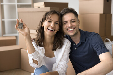 Happy attractive young couple sitting among cardboard moving boxes, holding key from new house, laughing, looking at camera with toothy smiles, celebrating relocation into new apartment, flat