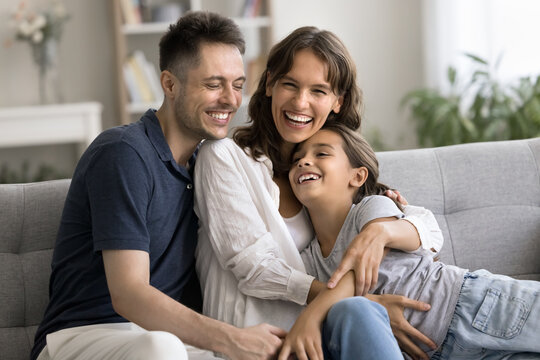 Excited happy young mother and father hugging little daughter kid on home sofa, tightening child with love, care, smiling, laughing, looking at camera, enjoying parenthood, leisure