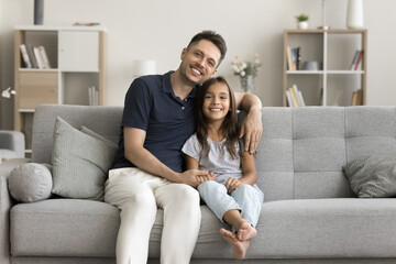 Obraz premium Happy handsome young dad and beautiful little daughter girl posing on comfortable sofa at cozy home, looking at camera, smiling, laughing, hugging. Father and kid family portrait