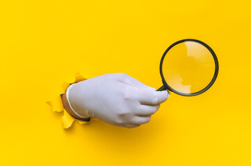 A hand in a white disposable glove holds a magnifying glass through a torn hole in yellow paper....
