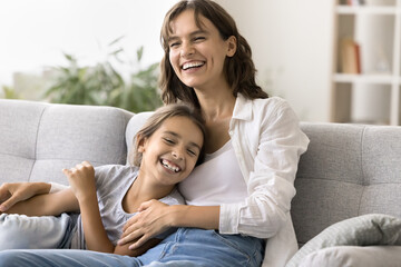 Cheerful young mom cuddling happy laughing daughter kid on home couch, looking away, laughing,...