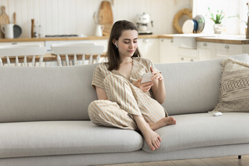 Young woman relaxing on cozy couch with modern smartphones, wear wireless earphones listen received...