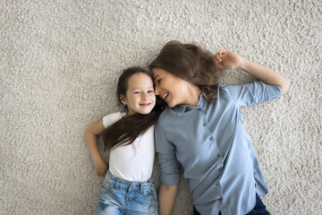 Above top view cute little girl with loving mother lying on soft carpet on warm floor with underfloor heat system smile look at camera, having fun enjoy carefree time together at home. Family, leisure