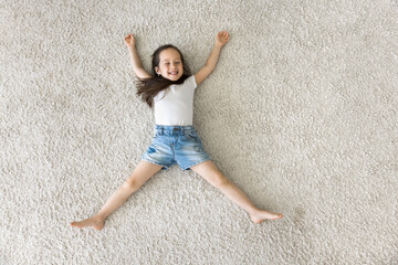 Above view playful beautiful little girl relaxing at home, spread her arms and legs lying on warm floor, looks carefree, feel happy, spend free time indoor, enjoy soft pile of new bought beige carpet