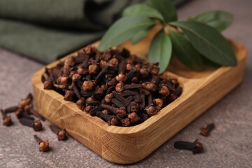 Wooden tray with aromatic cloves and green leaves on brown table, closeup