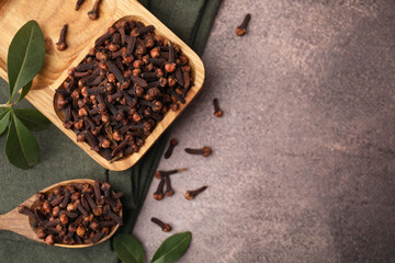 Wooden tray with aromatic cloves, spoon and green leaves on brown table, flat lay. Space for text