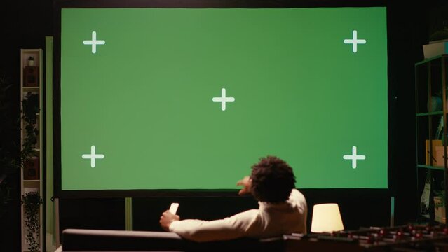 African american man using video projector to binge TV series on subscription based streaming services. Cord cutter enjoying VOD shows on gigantic green screen television set