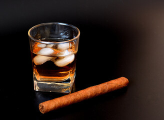 A glass of whiskey with ice cubes and a large Cuban cigar on a black background.