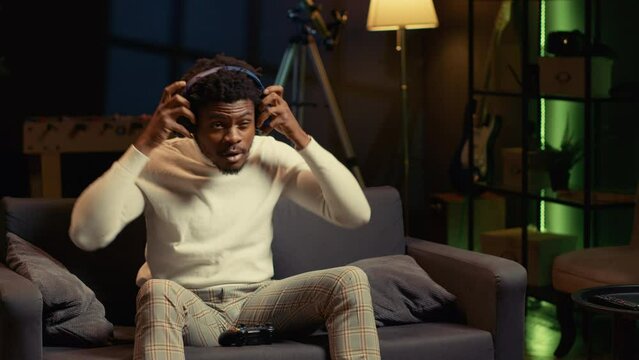 Man in rgb lit apartment playing video games on couch, enjoying day off from work. Gamer battling enemies in online multiplayer shooter on gaming console, talking with teammates through headphones