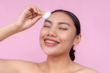 Smiling Asian woman using a cotton ball to apply toner on her forehead, promoting a clean and...