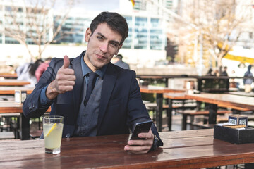 Businessman sitting at an outdoor bar drinking lemonade looking to the camera giving a thumb up - obrazy, fototapety, plakaty