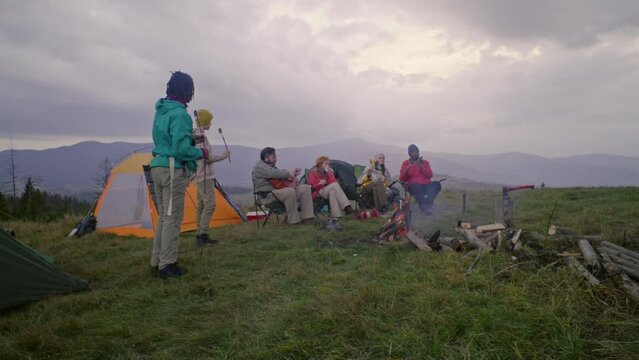 Diverse hiking buddies rest in camp on hill. Boys stand near campfire and eat marshmallow. Adult Caucasian man sits with guitar and talks with his friends. Tourists during vacation trip to mountains.