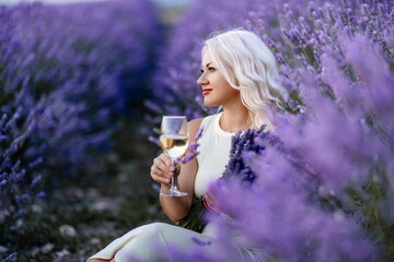 Blonde lavender field holds a glass of white wine in her hands. Happy woman in white dress enjoys lavender field picnic holding a large bouquet of lavender in her hands