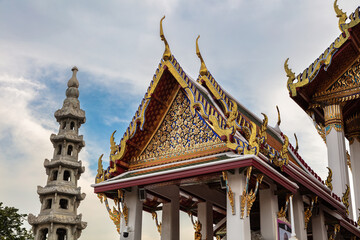 Decorated roof at Wat Suthat in Bangkok, Thailand. Intricate colored and golden ornamentation. Buildings and status on either side. Blue sky and clouds behind. 
