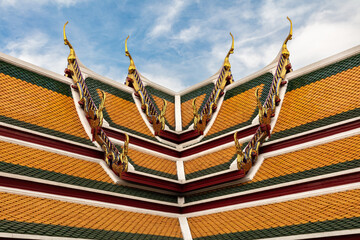 Fototapeta na wymiar Decorated roof at Wat Suthat in Bangkok, Thailand. Design of orange, red and green tiles, golden ornamentation. Blue sky and clouds beyond. 