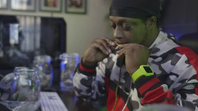 side view of a black man with a black head wrap is licking a blunt to seal the marijuana into the tobacco wrapper with mason jars of cannabis buds