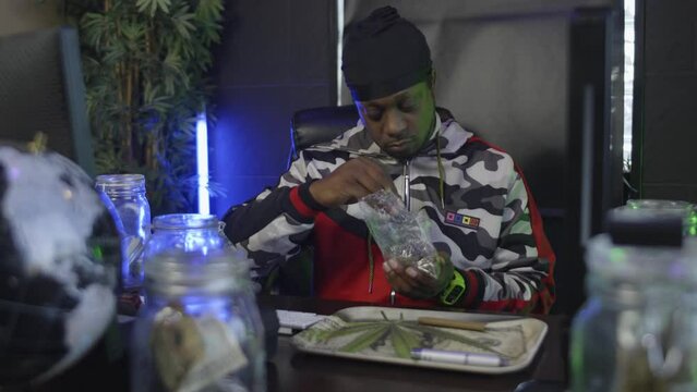 straight on view of a black man reaching into a bag of marijuana and removing buds which he breaks up onto a pot leaf rolling tray with a mason jar of money  and a globe on the desk in front of him