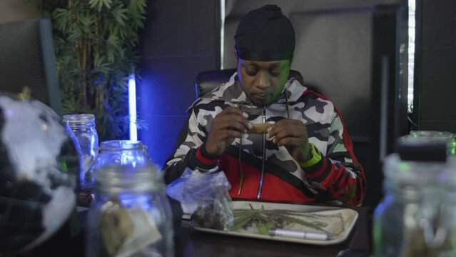 straight on view of a black man licking a blunt to seal to tobacco wrapper around the weed there is a rolling tray on the desk  and a jar of money in the foreground