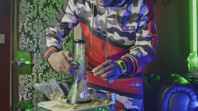 A black man lights a wick which he utilizes to light a marijuana water bong and takes a toke of pot or weed cannabis