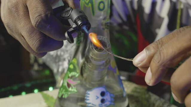 a black mans hand using a torch to light a wick in preparation of smoking a water bong filled with marijuana