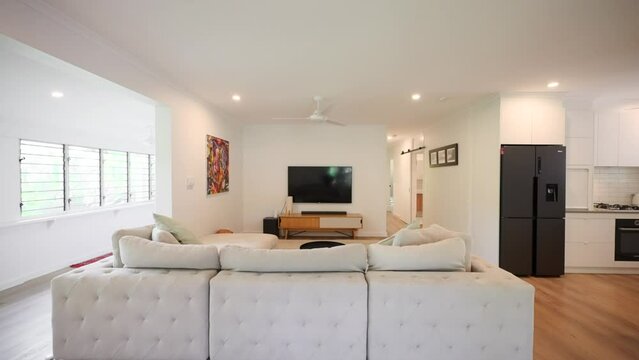Modern lounge room with warm tones, white sofa and louver windows letting in natural light. Kitchen adjacent with black fridge visible in frame in interior of home house.