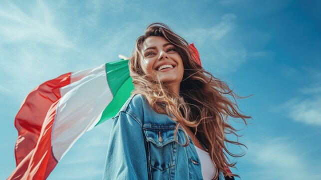 Happy young long haired woman in denim jacket holding italian flag against blue sky
