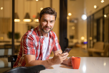 Man looking at phone at home, browsing mobile apps, reading news, chatting or shopping online, holding smartphone. Attractive man in casual clothes at the living room, talking on mobile phone.