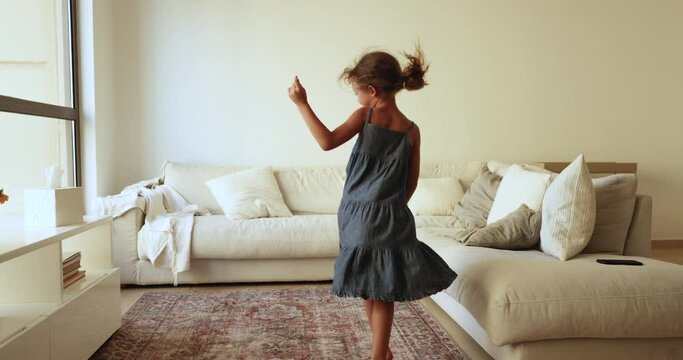Cute little girl in casual summer dress sings a song and dancing in modern living room, lively moving, jumping, looking happy and untroubled. Funny adorable preschooler child having fun alone at home