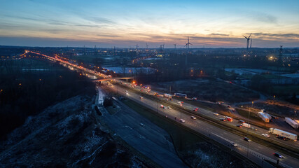 This image captures the dynamic scene of twilight on the E19 near Halle, with the rush of traffic juxtaposed against a backdrop of stationary wind turbines. The contrast between the movement on the