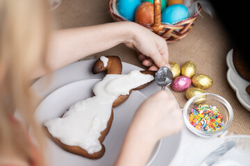 Unrecognizable blond child topping Easter bunny shaped cookie with icing on plate. Multi colored...