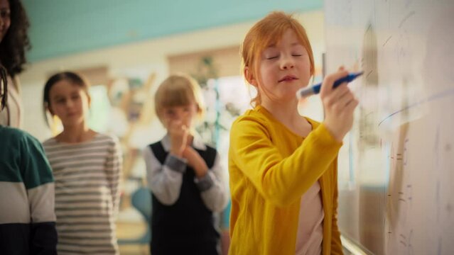 Smart Little Girl Solving an Equation in Front of a Whole Class and Teacher in Primary School. Young Attentive Female Writing Down the Answer on a Board, Happy to Provide a Solution to an Exercise