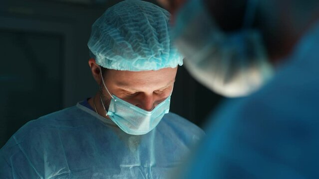 Closeup face of focused serious male surgeon wearing surgical cap and mask processing surgical operation in dark operating room of modern hospital emergency department. Shooting in slow motion.