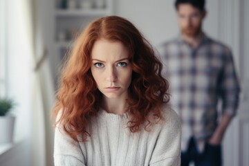 Sad young woman with curly red hair and man argument, сlose up upset wife in front and husband standing behind quarrel at home. Family conflict, crisis, psychological abuse, relationships concept