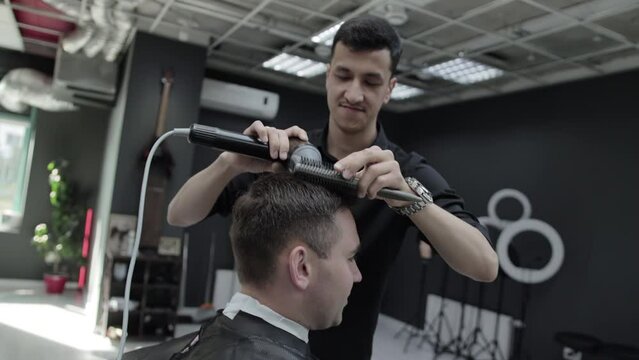 Handsome male brunette hairdresser styles the hair of a male client with a hairdryer and comb in a barbershop. Slow motion