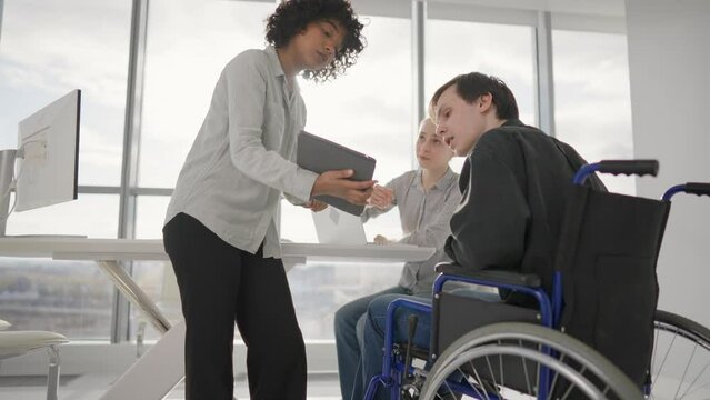 Paralyzed man works with colleagues. Guy in wheelchair has spine trauma discussing working project in office on meeting with multiethnic team. Teamwork brainstorming, special possibilities needs.